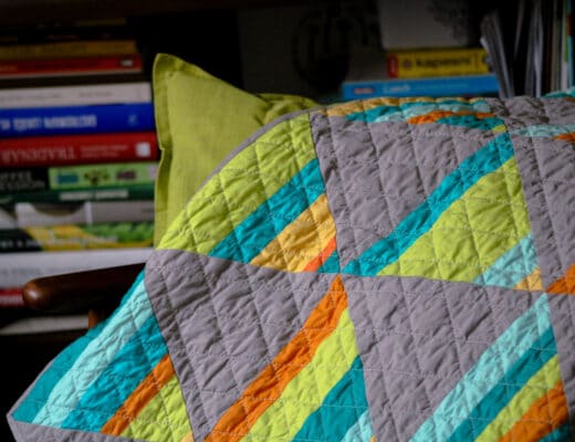Barevná prošívaná patchworková deka leží přehozená přes zelený polštář. V pozadí jsou rozmazané knihy.